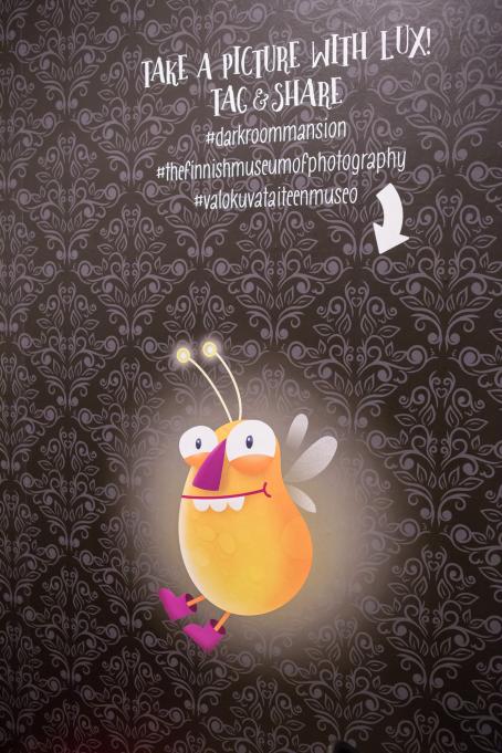 Tummalla tapetilla peitetyllä seinällä on kuva oranssista, hohtavasta ötökästä ja yläpuolella teksti: "Take a picture with Lux! Tag and share #darkroommansion "thefinnishmuseumofphotography #valokuvataiteen museo".