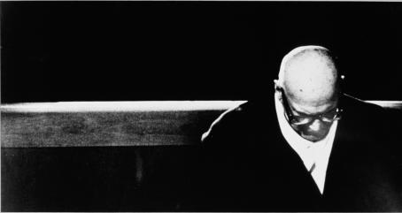 Mustavalkoisessa kuvassa Urho Kekkonen istuu yksin kirkonpenkissä pää painuksissa. Kuvassa on vahva kontrasti, tausta on aivan musta. 