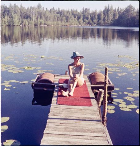 Nainen istuu laiturilla punaisen viltin päällä. Hänellä on sininen lierihattu päässä. Vedessä on lumpeenlehtiä ja aurinko paistaa.