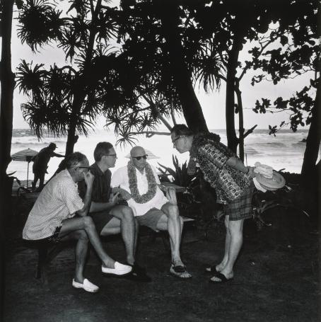 Mustavalkoisessa kuvassa neljä miestä ulkona merenrannalla, palmupuiden tms. alla. Kolme miehistä istuu ja neljäs seisoo heidän edessään kumartuneena eteenpäin. Kaikilla on shortsit ja lyhythihaiset kauluspaidat. Kahdella miehellä on kukkaseppele kaulassa. 