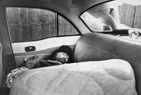 Mustavalkoisessa kuvassa mies makaa nukkumassa auton takapenkillä. Hänellä on karvalakki päässä. Auton takalasissa on iso reikä. Auton ulkopuolella näkyy lauta-aitaa.