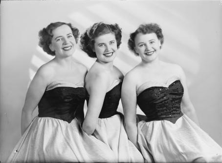 Mustavalkoisessa kuvassa vierekkäin kolme leveästi hymyilevää naista. Kaikilla on päällään mekko, jossa on tumma, korsettimainen yläosa ja vaalea hameosa. Kaikilla on melko lyhyet, laineikkaat hiukset. 