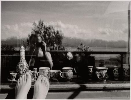Mustavalkoinen omakuva, jossa kuvaaja heijastuu epäselvästi ikkunasta tms. Kuvaajan takana näkyy järvi ja tunturimaisemaa. Ikkunan toisella puolella on koristeellisia kahvikuppeja ja muita astioita. Kuvaajan jalkaterät näkyvät ikkunankarmia vasten. 