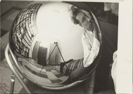 Mustavalkoisessa kuvassa pallon muotoinen heijastava esine, josta heijastuu miehen kasvot ja hänen takanaan oleva huone. Huoneessa on ainakin sänky ja leveä kirjahylly. 