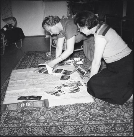 Mustavalkoisessa kuvassa kaksi naista istuu lattialla. He ovat kumartuneet lattialle levitetyn kartan ja sen päällä olevien valokuvien ylle. 