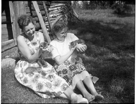 Mustavalkoisessa kuvassa kaksi naista istuu nurmikolla kesämekoissa. Molemmilla on sylissä ja käsissä kissanpentuja. Molemmat naiset hymyilevät. Aurinko paistaa. 
