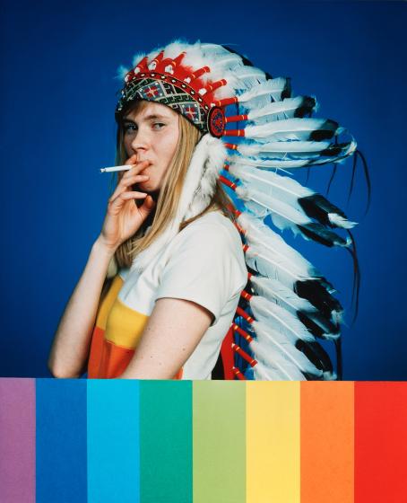 Nuorella naisella on päässään sulkapäähine. Hän polttaa tupakkaa ja katsoo sivusilmällä kameraan. Kuvan alareunassa on sateenkaaren värit pystyviivoina. 