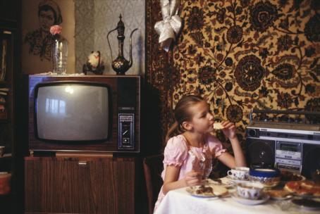 Nuori tyttö vaaleanpunaisessa mekossa istuu syömässä pienen pöydän ääressä. Pöydällä on iso vanha kasettisoitin seinää vasten. Seinää peittää iso, ruskeansävyinen, koristeellinen kangas. Tytön takana on putkitelevisio. 