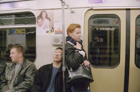 Punahiuksinen nainen seisoo metrossa ovien vieressä pitäen tiukasti käsilaukustaan kiinni. Naisen selän takana penkillä istuu kaksi miestä. 
