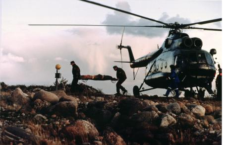Kivisessä maastossa on helikopteri laskeutuneena. Sen vieressä kaksi henkilöä kantaa paareilla kolmatta henkilöä poispäin helikopterista.