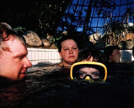 Ihmisiä uima-altaassa. Kuva on otettu lähes vedenpinnan tasolta. Lähellä altaan reunaa on kaksi aikuista kaulaa myöten vedessä. Heidän edessään on lapsi sukellusmaski päässä, nenää myöten vedessä. Altaan reunan ulkopuolella on kiviä ja viherkasveja. 