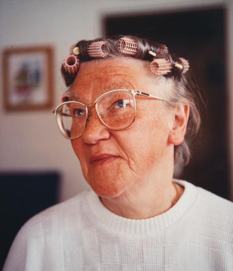 Muotokuva vanhasta naisesta. Naisella on valkoinen neule, isot vaaleat silmälasit ja hiuksissa papiljotteja. Hänen katseensa kohdistuu yläviistoon. 