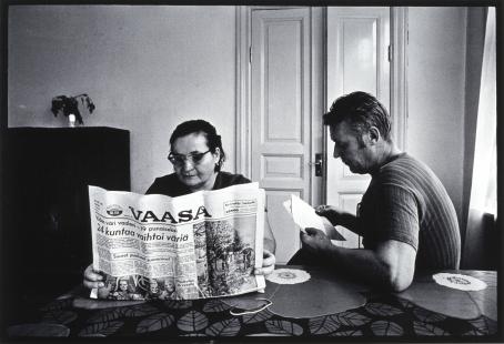 Mustavalkoisessa kuvassa nainen ja mies istuvat pöydän ääressä. Nainen lukee Vaasa-sanomalehteä. Isoin otsikko on "24 kuntaa vaihtoi väriä". Vieressä mies lukee jotain paperia, ehkä laskua tms. Pöydällä on lehtikuvioinen liina ja kukan malliset tabletit. 