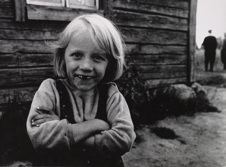 Mustavalkoisessa kuvassa vaaleahiuksinen lapsi seisoo ulkona puisen talon edustalla. Lapsella on kädet puuskassa, hän hymyilee ja katsoo suoraan kameraan. Häneltä puuttuu kaksi etuhammasta. 