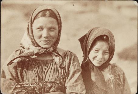 Mustavalkoisessa seepiansävyisessä kuvassa kaksi nuorta huivipäistä tyttöä. Molemmat hymyilevät ja katsovat jonnekkin kuvan ulkopuolelle. 