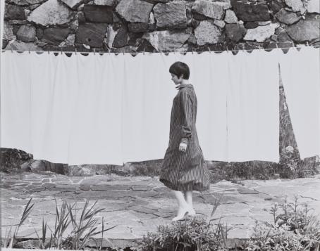 Mustavalkoisessa kuvassa lyhythiuksinen nainen raidallisessa mekossa kävelee pyykkinarulla roikkuvien lakanoiden edessä. 