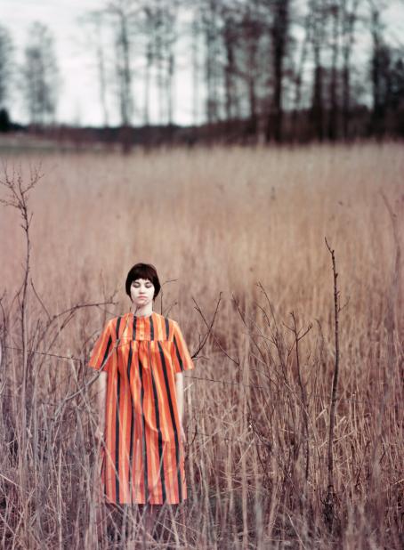 Lyhythiuksinen nainen oranssissa pystyraitaisessa mekossa seisoo niityllä tai pellolla. Naisella on silmät kiinni. Hänen ympärillään kasvaa vaaleanruskeaa heinää. 