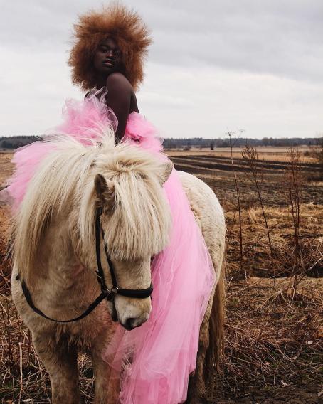 Tummaihoinen nainen istuu pienen hevosen tai ponin selässä. Hänellä on päällään vaaleanpunainen tyllihame tai -mekko, jonka helma roikkuu ponin kylkeä vasten lähes maassa asti. Naisella on vaaleanruskea afrotukka. Takana näkyy kynnettyä peltomaisemaa.