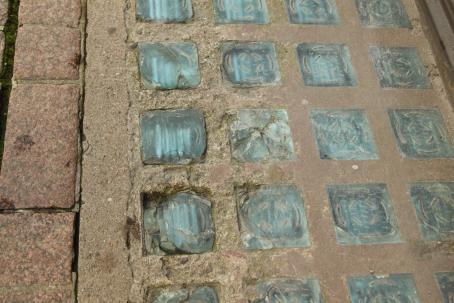 Maassa betoniin upotettuna vaaleansinisen sävyisiä neliöitä riveissä. Osa neliöistä on rikkoutunut ja lohjennut.