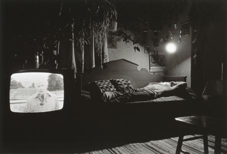 Mustavalkoisessa kuvassa mies makaa puisella sivustavedettävällä sohvasägyllä. Sängyn yläpuolella oleva lamppu valaisee huoneen. Sängyn vieressä on televisio, jonka ruudulla on mies ja teksti "Aarne Tanninen, Washington". 