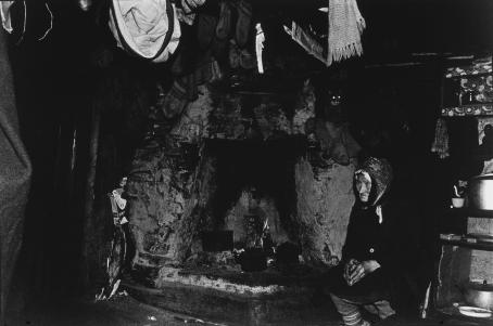 Mustavalkoisessa, tummasävyisessä kuvassa massiivisen ja likaisen tulisijan vieressä istuu vanha nainen tummissa vaatteissa. Ylempänä takan reunalla istuu kissa, josta erottuu lähinnä vain kiiluvat silmät. Kuvan yläreunassa näkyy osittain katosta roikkuvia vaatteita. 