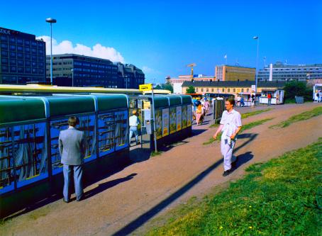 Harmaatakkinen mies seisoo bussipysäkin takana katse pysäkin seinää päin, selkä kameraan. Kuvan oikeassa alareunassa on nurmikkoa ja sorapolku, jota pitkin kävelee valkopaitainen ihminen pysäkkiä kohti. Aurinko paistaa ja taivas on sininen. 