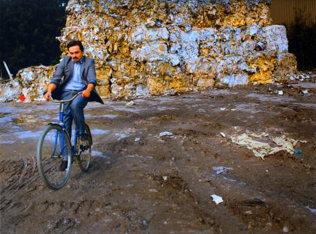 Harmaatakkinen, viiksekäs mies istuu sinisen pyörän selässä. Takana on roskia puristettuna isoiksi kuutioiksi ja kasattuna korkeiksi pinoiksi. 
