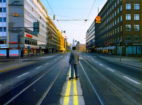 Harmaatakkinen mies seisoo skeittilaudan päällä keskellä tietä selkä kameraan päin. Tiessä on kahdet raitiovaunukiskot. Kadun molemmilla puolilla on kerrostaloja, kauempana olevia taloja valaisee aurinko. 