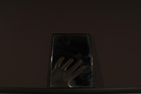 Tumman kuvan keskellä on suorakulmion muotoinen peili, josta heijastuu epätarkkana ihmisen käsi sormet levällään.