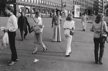 Mustavalkoisessa kuvassa kävelee ihmisiä aukiolla eri suuntiin. Keskelle on pysähtynyt nuori tummaihoinen mies leveälahkeisissa housuissa. Taustalla näkyy Kansallis-osake-pankin rakennus.
