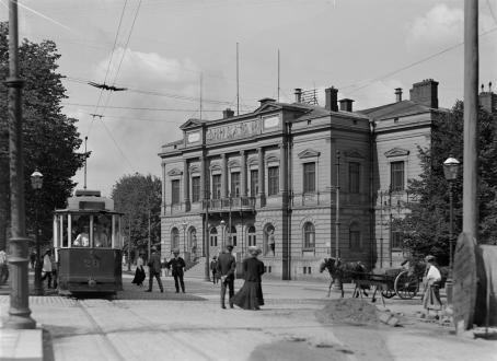 Mustavalkoinen vanha maisema Helsingistä. Vasemmalla on raitiovaunu, kadulla kävelee ihmisiä, siellä on myös hevoskärryt ja taustalla on koristeellinen rakennus, joka on Ylioppilastalo. 
