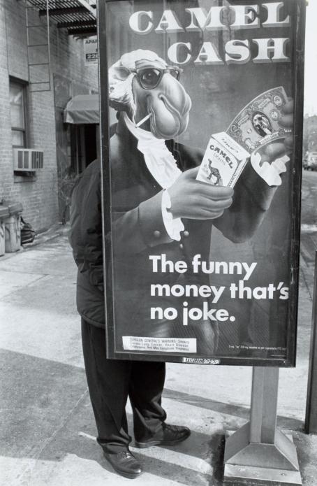 Mustavalkoisessa kuvassa kadulla oleva mainos, jossa on ihmismäiseksi puettu kameli. Mainoksessa lukee "Camel Cash, the funny money that's no joke". Mainoksen takana seisoo henkilö, joka on osittain piilossa mainoksen takana ja sulautuu mainoksessa olevaan kameliin. 