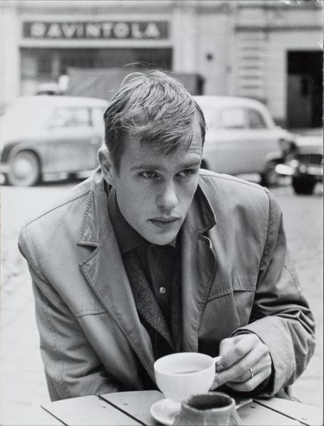 Mustavalkoisessa kuvassa nuori, nahkatakkinen mies istuu ulkona ja pitelee kahvikuppia. 