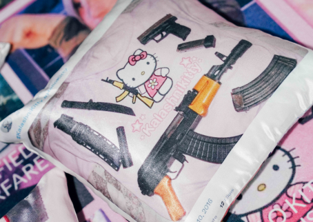 Tyyny, jossa rynnäkkökivääriä pitelevä Hello Kitty -hahmo. Hahmon ympärillä erilaisia aseita. Tyynyn taustalla videopelihahmo ja vaaleanpunaista söpöä estetiikkaa.