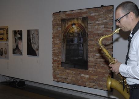 Näyttelyn kuvia esillä näyttelytilassa. Mies soittaa saksofonia kuvien edessä. 