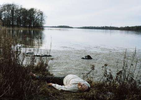 Henkilö makaa selällään järven rannalla rantakasvillisuuden keskellä.