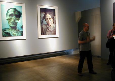 Etualalla seisoo valokuvaaja Mario Rizzi. Hänen takanaan seinällä näkyy kaksi hänen ottamaansa kuvaa muumioista. 