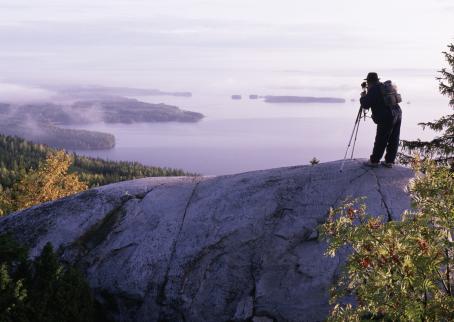 Maisema, jossa etualalla kalliota, taustalla sumuista metsää ja järvi. Kallion päällä on valokuvaaja ottamassa kuvaa. 