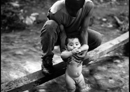 Mustavalkoisessa kuvassa kommandopipoinen henkilö uittaa alastonta poikavauvaa virtaavassa purossa. 