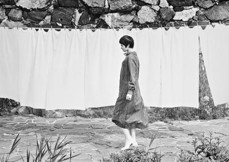 Mustavalkoisessa kuvassa lyhythiuksinen nainen raidallisessa mekossa kävelee pyykkinarulla roikkuvien lakanoiden edessä. 