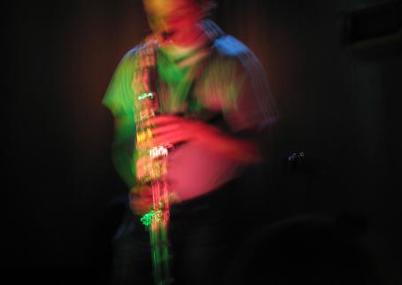Hieman tärähtäneessä kuvassa henkilö soittaa saksofonia. Kuvassa näkyy vihreän ja vaaleanpunaisen sävyjä.