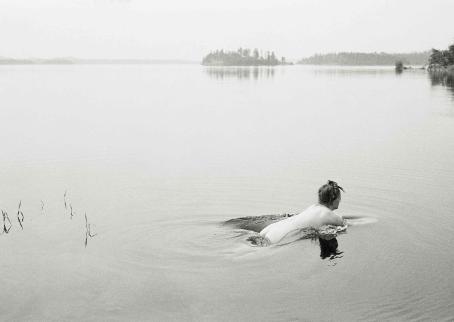 Mustavalkoinen järvimaisema, jossa alaston nainen on juuri kastautumassa veteen.