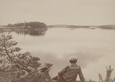 Mustavalkoisen kuvan etualalla on kaksi henkilöä katselemassa järvimaisemaa. 