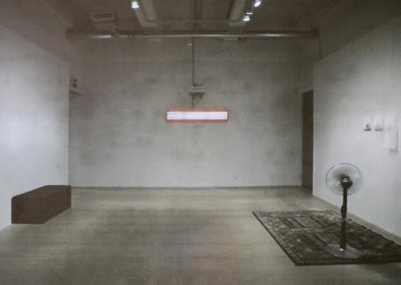 Lähes tyhjä huone. Vasemmalla seinustalla on ruskea suorakulmion muotoinen penkki, takaseinällä on pitkänomainen lamppu ja oikealla seinustalla on maton päällä tuuletin ja seinällä papereita.