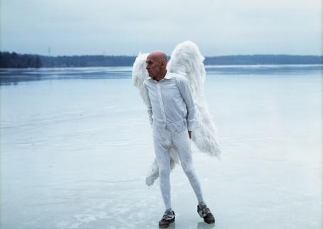 Vanha mies valkoisissa vaatteissa ja valkoiset enkelinsiivet selässään seisoo järven jäällä sinisävyisessä maisemassa.