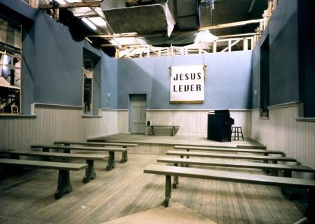 Elokuvan lavasteeksi tehty huone. Kuvan etualalla on penkkejä rivissä, huoneen etuosassa on piano ja seinällä roikkuu kangas jossa lukee "Jesus lever".