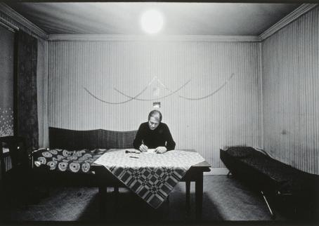 Mustavalkoinen kuva huoneesta. Huoneen keskellä on pöytä, jonka ääressä henkilö istuu kirjoittamassa paperille. 