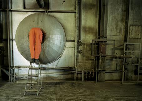 Näkymä tehtaasta. Vasemmassa reunassa oranssihaalarinen henkilö seisoo jakkaran päällä kurkistamassa putkeen, ja hänestä näkyvät vain jalat.