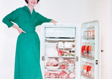 Nainen vihreässä mekossa seisoo häntä lyhyemmän jääkaapin vieressä nojaten siihen. Jääkaapin ovi on auki, ja se on täynnä ruokaa.