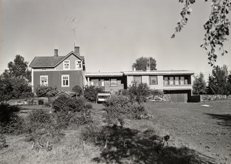 Mustavalkoisessa kuvassa etualalla pihaa, pihan takana kaksi taloa. Vasemmalla vanhempi puutalo, jonka vieressä uudempi matala tiilitalo. 